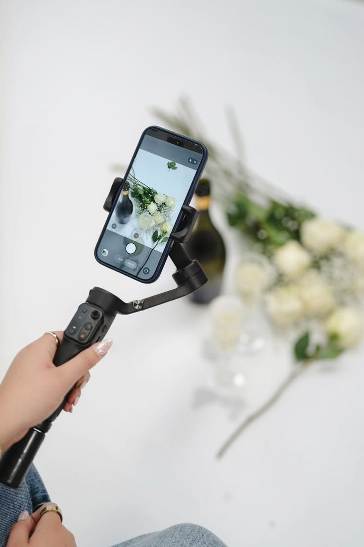 A hand holds a smartphone gimbal stabilizer photographing white roses and a bottle on a white background.
