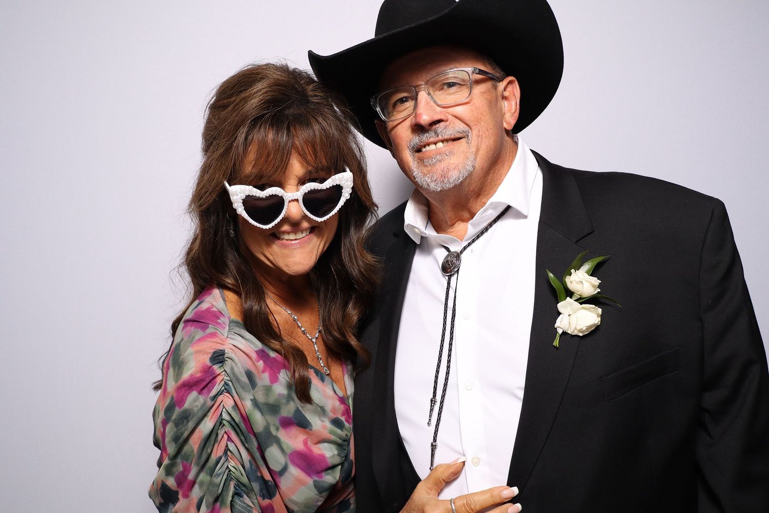 Couple posing in photo booth, woman wearing heart-shaped novelty glasses, man in black cowboy hat and suit with boutonniere.