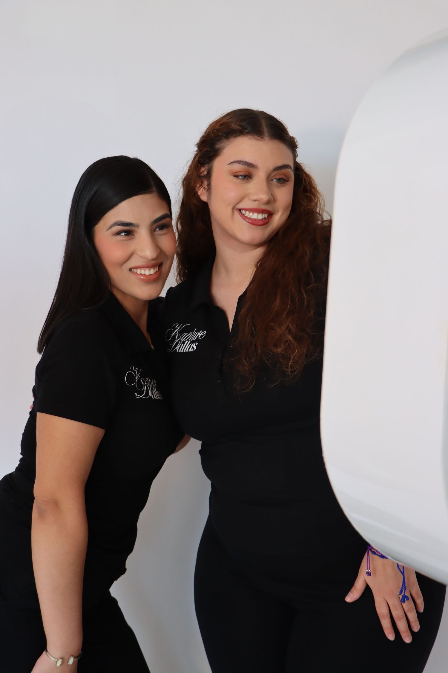 Two smiling women in matching black uniforms pose together in a bright white studio setting.