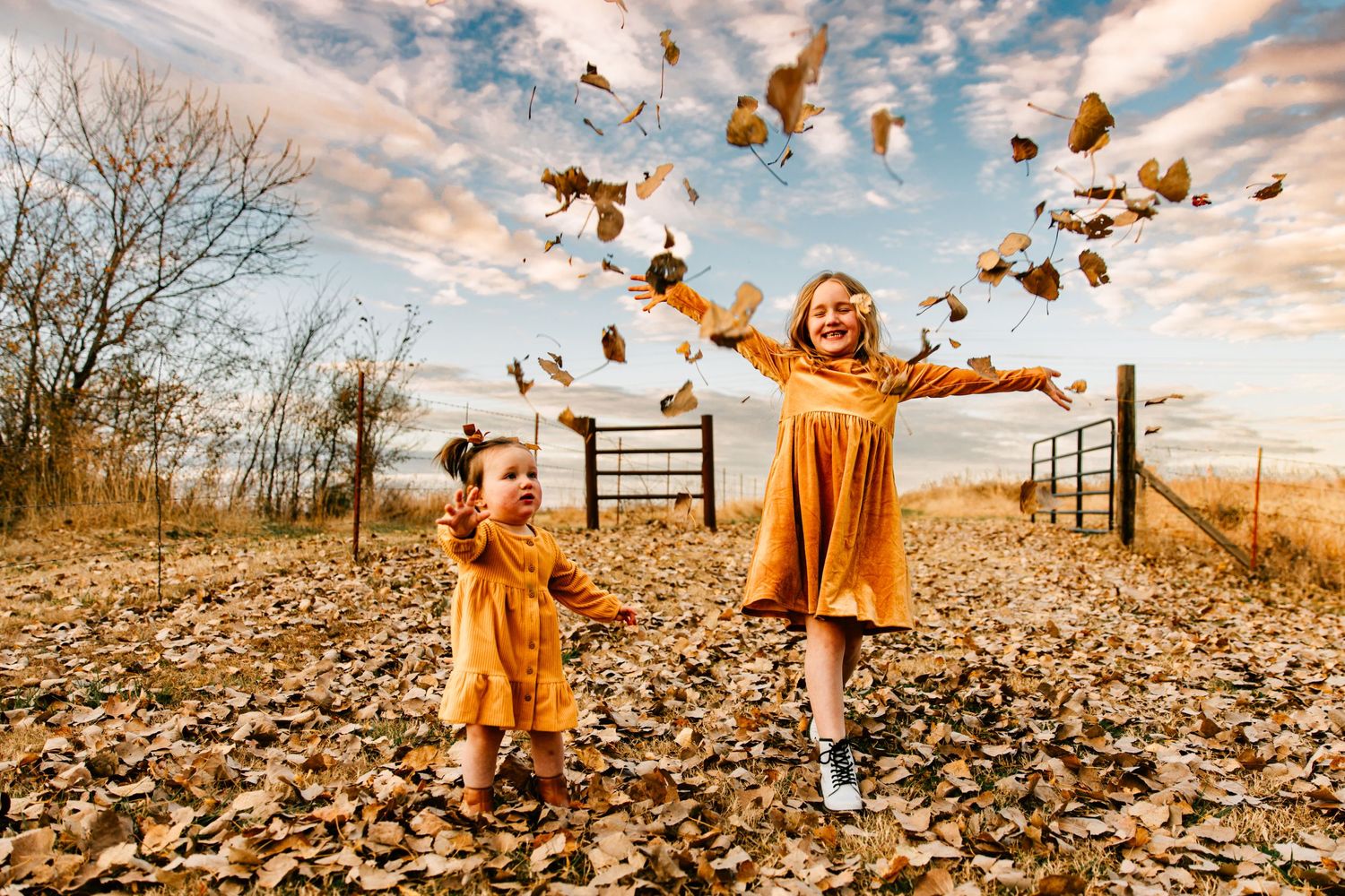 A bold, true-to-color photo of two young sisters throwing fall leaves at sunset in Kiefer, Oklahoma at Ogie Farms.