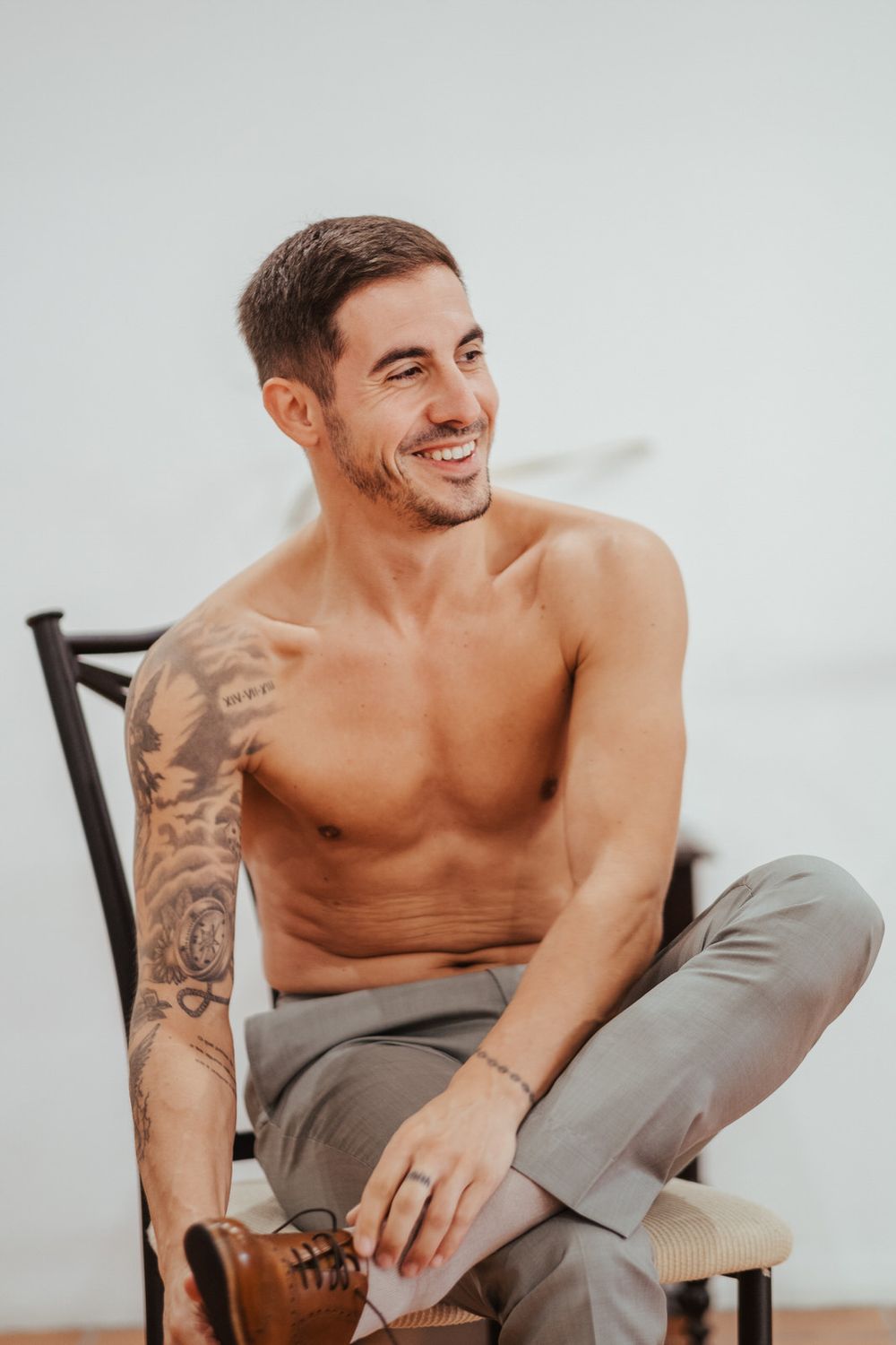 Smiling shirtless man with tattoos sitting on a chair wearing gray pants and brown shoes against a white background.