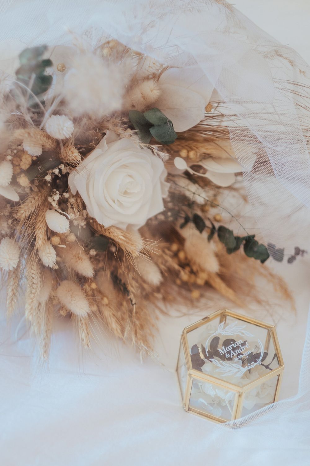 Elegant bridal bouquet with white roses, dried pampas grass, and a gold geometric glass ring box on white fabric.