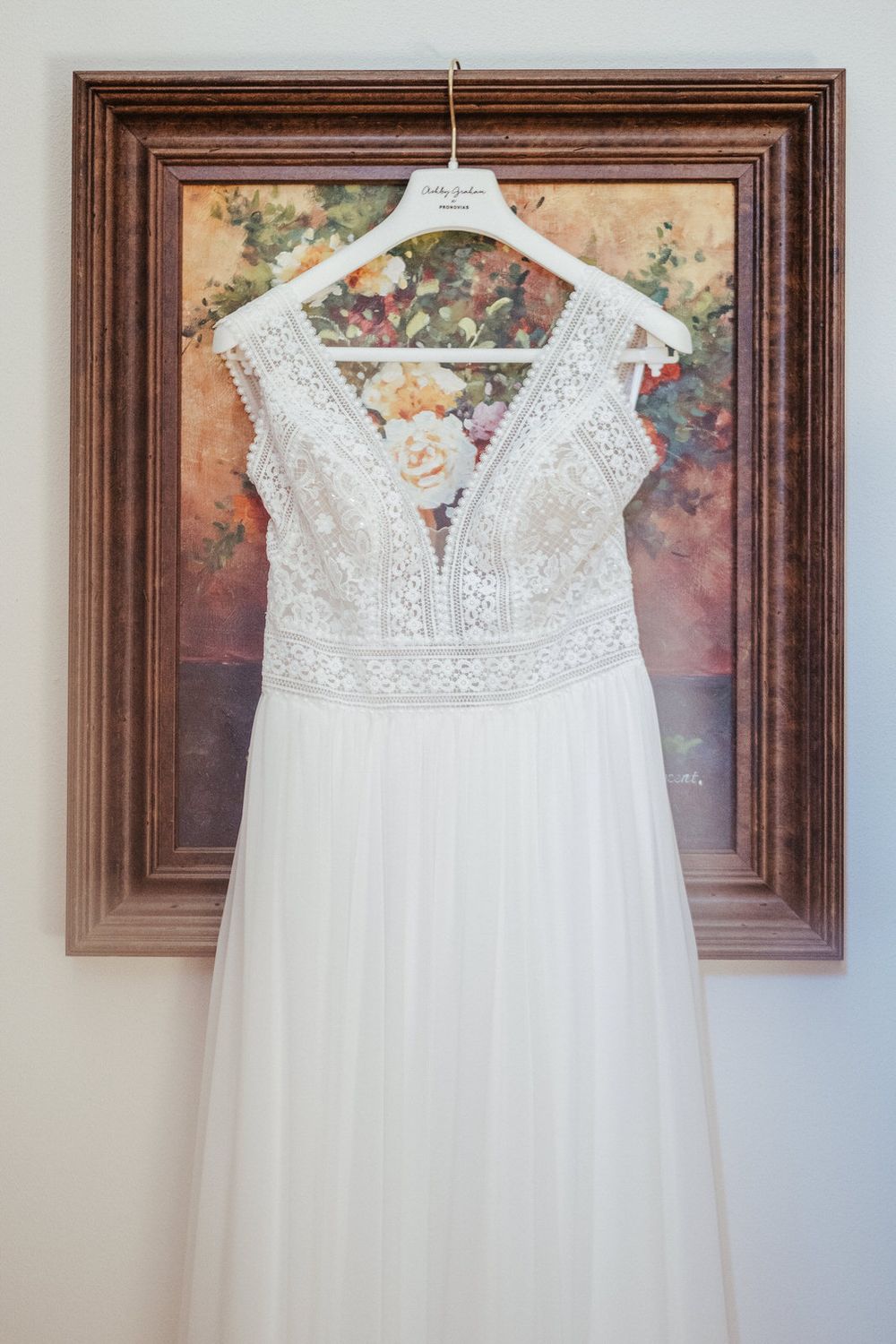 White lace wedding dress hanging on a decorative hanger in front of a floral painting in an ornate wooden frame.