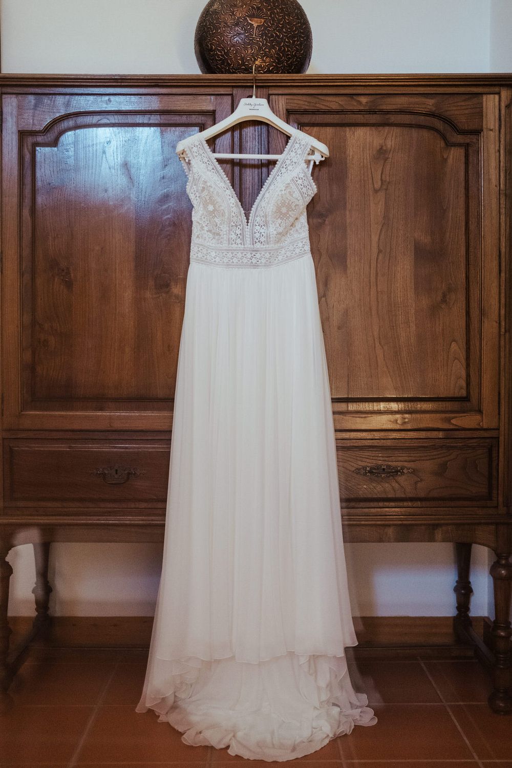 Elegant white lace bodice wedding dress hanging on wooden hanger against rustic wood armoire cabinet.
