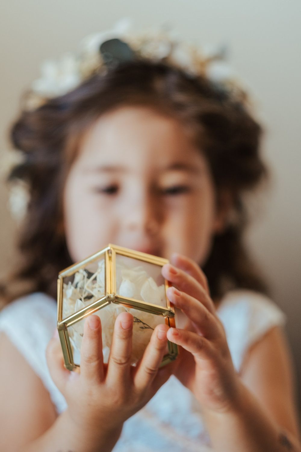 Young flower girl with curly hair and floral crown holds a gold geometric glass ring box at a wedding.