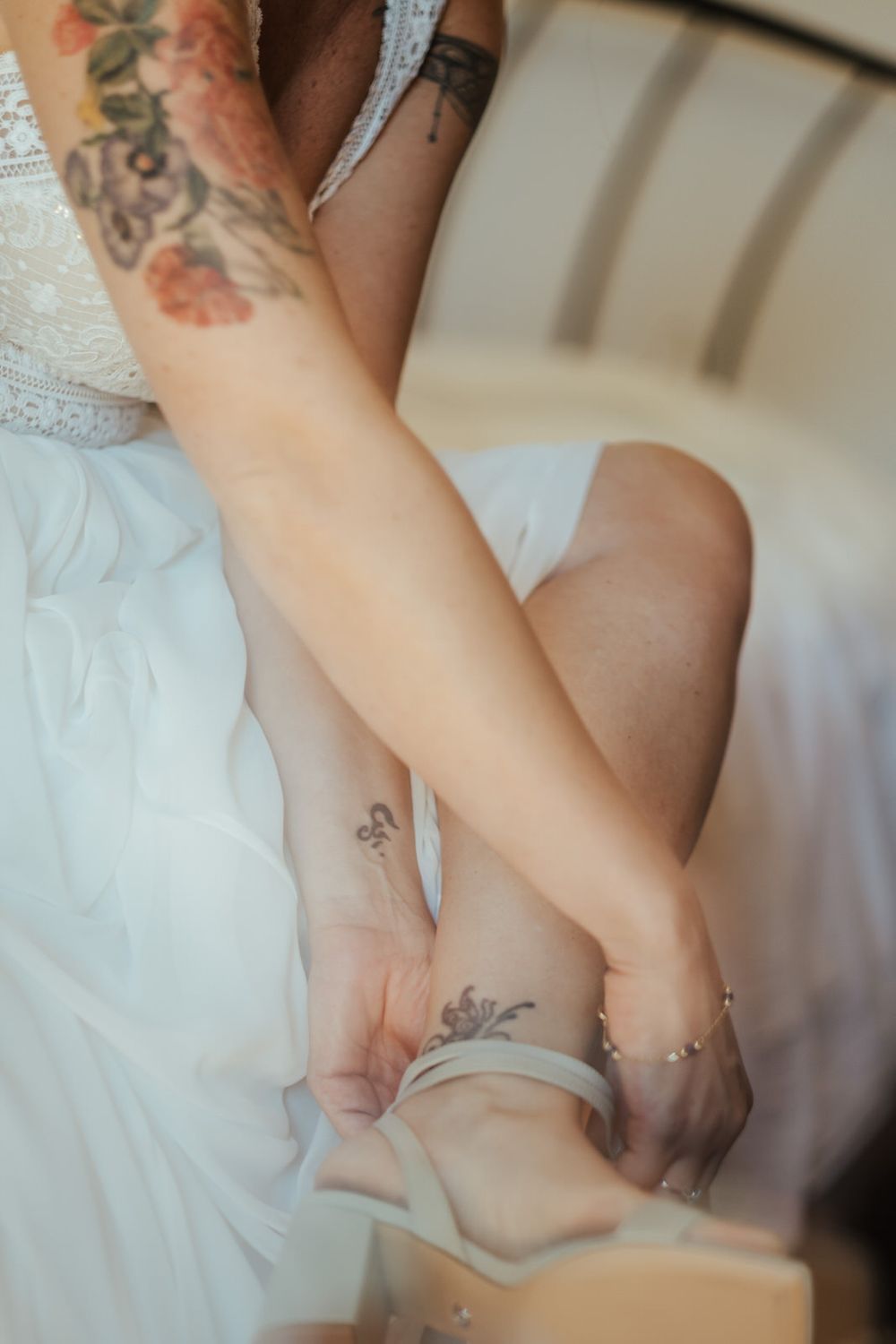 Tattooed bride in white dress fastening a delicate ankle strap shoe, with floral arm tattoos visible.
