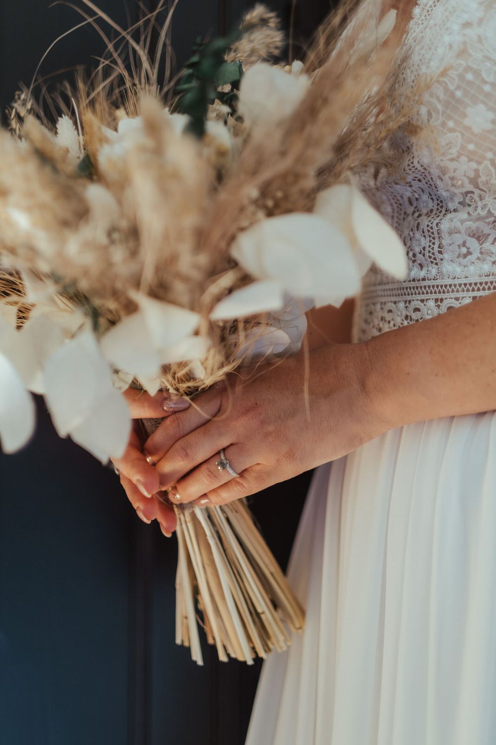 Bride in white lace dress holding a boho dried flower bouquet with pampas grass, white blooms, and a diamond ring.