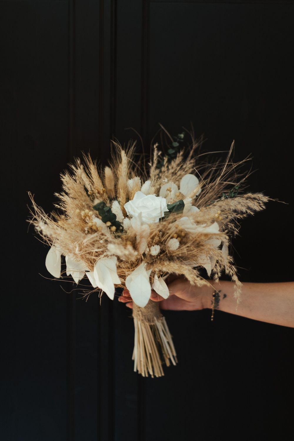 Hand holding a rustic boho bridal bouquet with dried pampas grass, wheat, and white roses on black background.