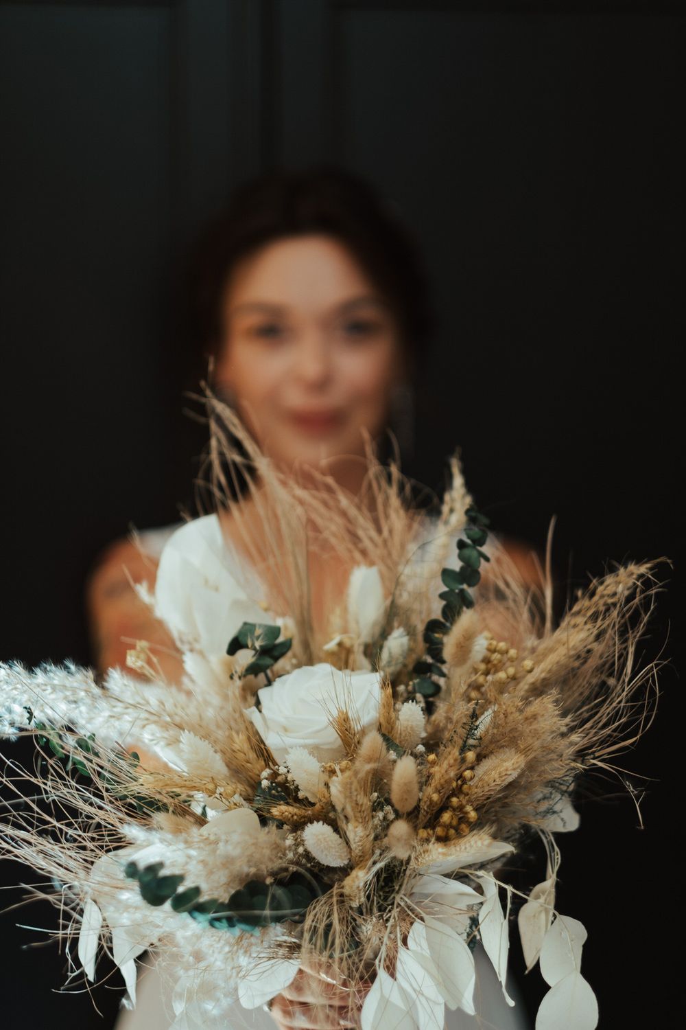 A bride in white holds a boho dried flower bouquet with pampas grass, wheat, and white roses toward the camera.