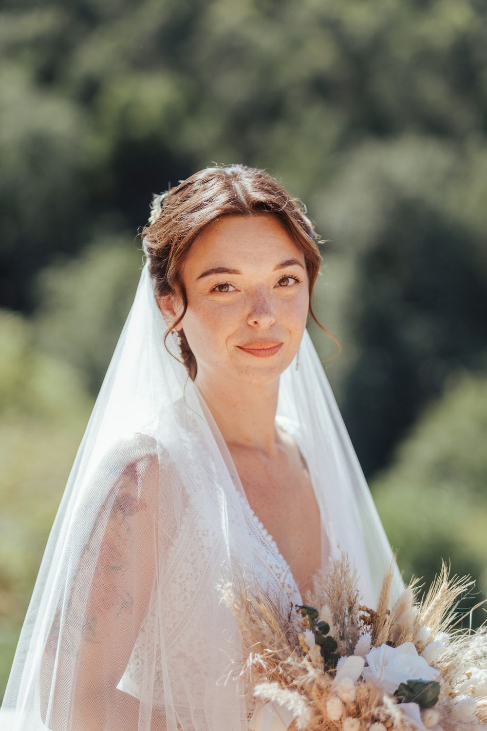 A radiant bride in a white lace gown and veil holds a boho dried flower bouquet outdoors in natural sunlight.