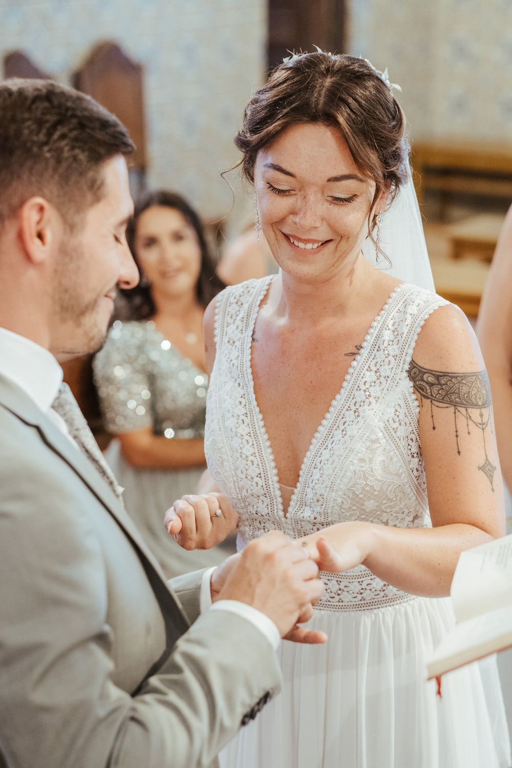 Bride smiling as groom places wedding ring on her finger during indoor ceremony, wearing white beaded dress with veil.