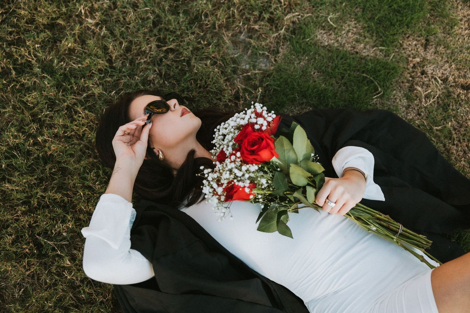 Person lying on grass wearing sunglasses and black blazer holding a bouquet of red roses and white flowers.
