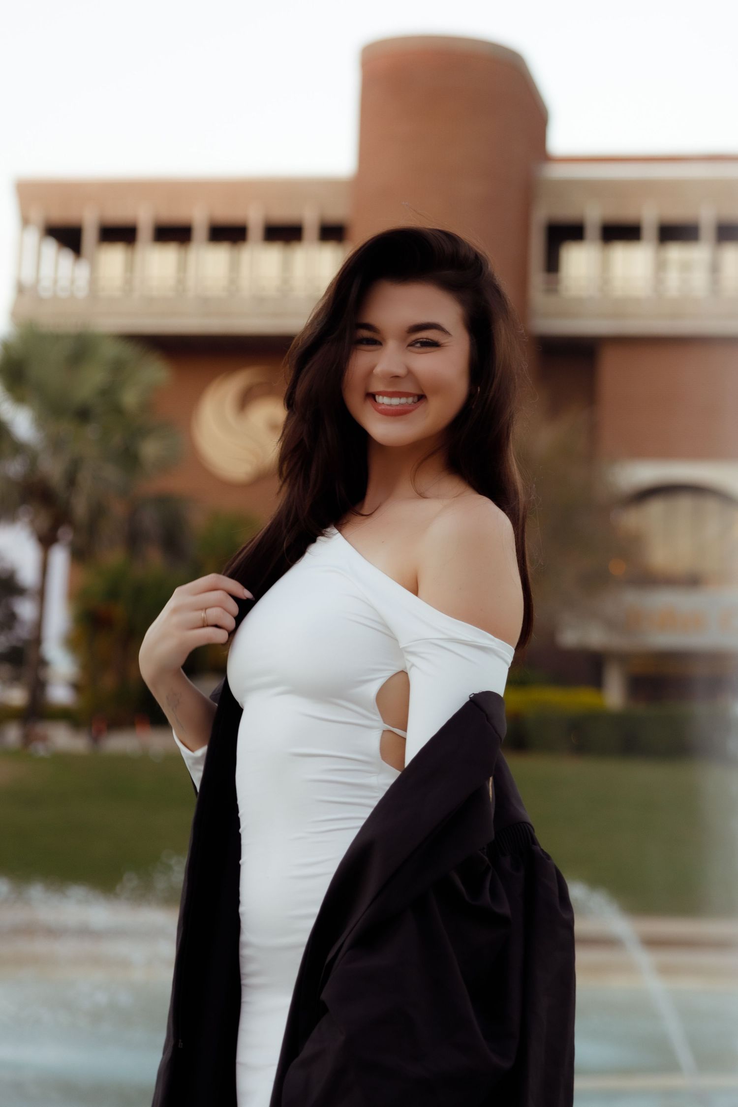 Young woman smiling in white off-shoulder dress with black jacket, posing outdoors near a fountain and building.