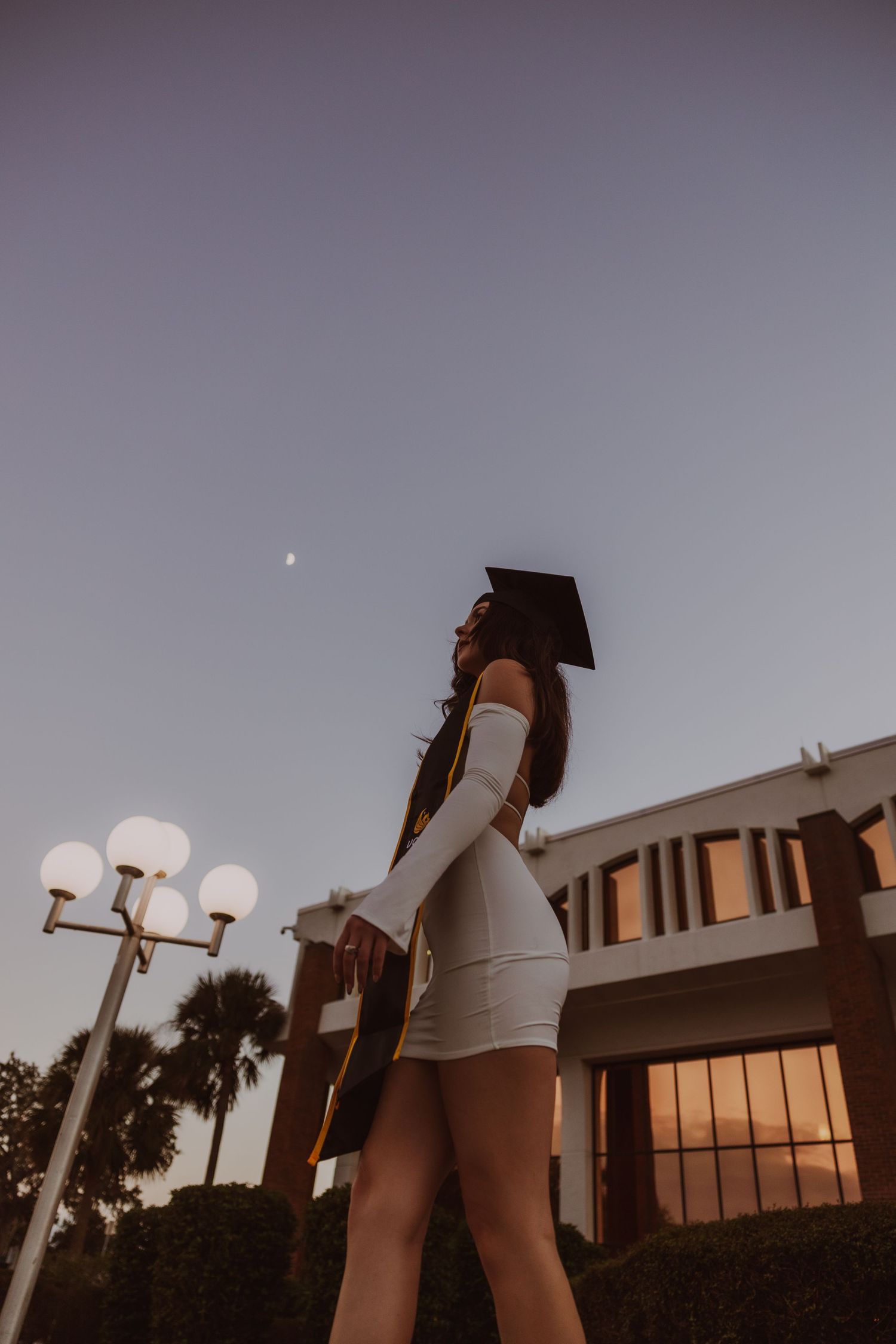 Young woman in white dress and graduation cap poses outdoors at dusk near a university building.