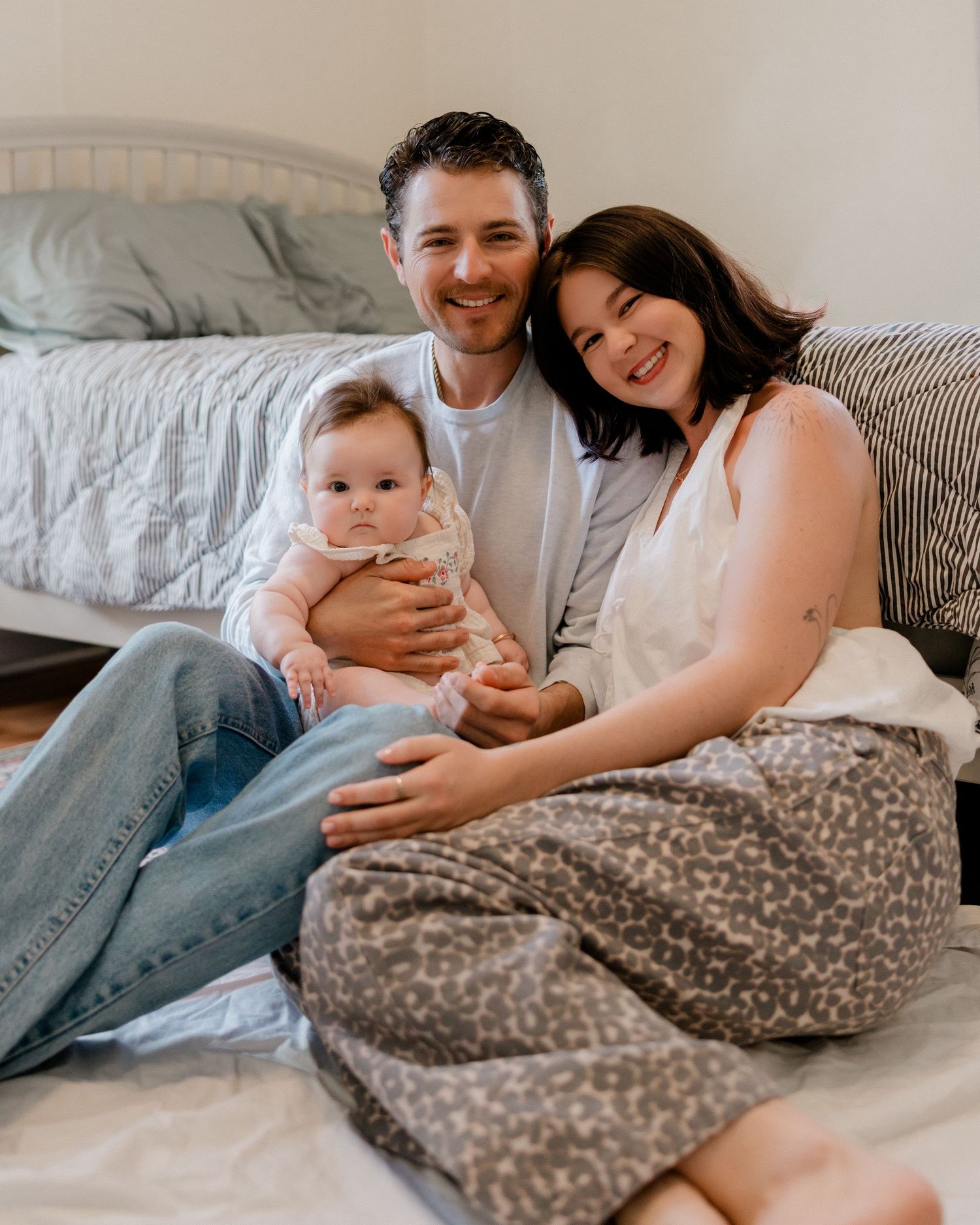 Happy couple sitting on bed smiling with their baby girl in a cozy bedroom setting.
