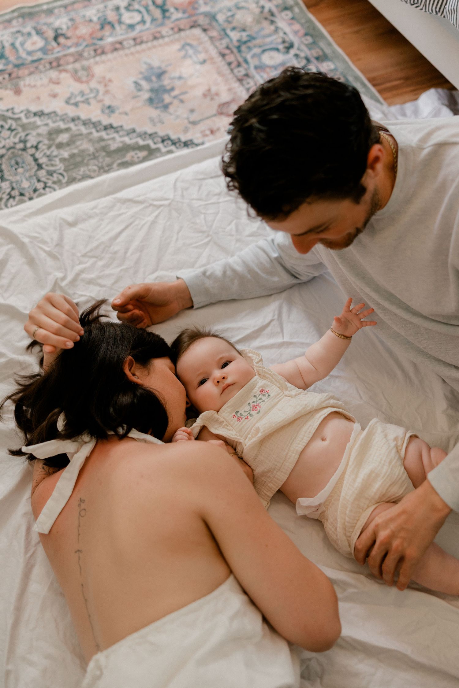 Parents lying on white bed with their baby, sharing a tender family moment at home.
