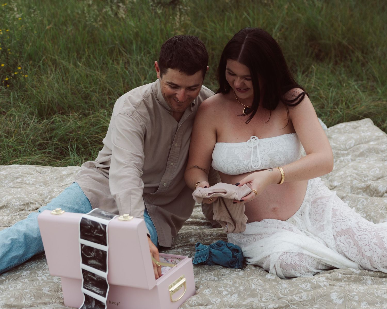 Expecting couple sitting on blanket outdoors, smiling while looking at baby ultrasound photos from a pink keepsake box.