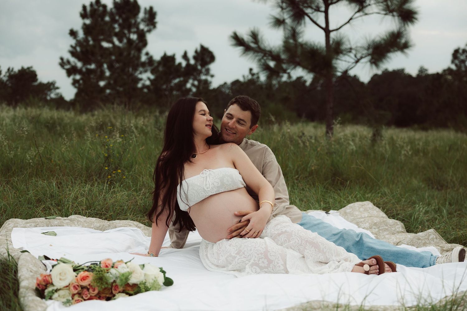 Couple sharing a tender maternity photo on a white blanket in a lush green field, woman in white dress cradling baby bump.