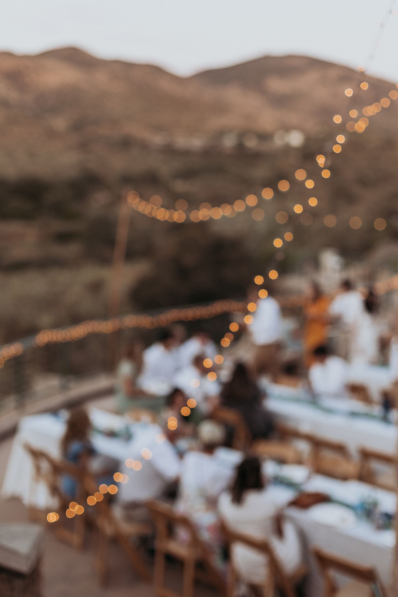 Blurred outdoor evening gathering with string lights, guests in white attire, and desert hills at sunset at Mission Trails Community Center.