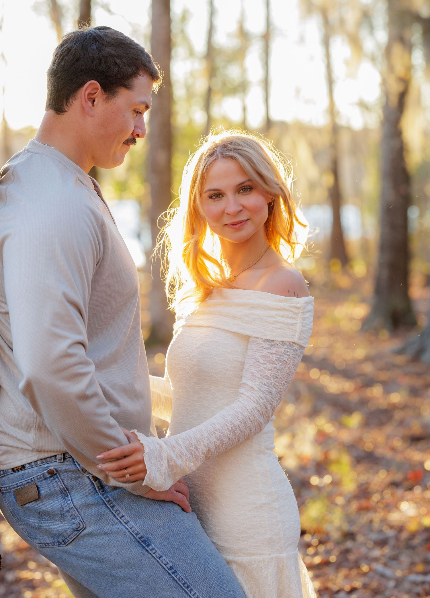 Couple in autumn forest, woman in white off-shoulder gown smiling at camera, man in jeans and beige shirt, golden sunlight.