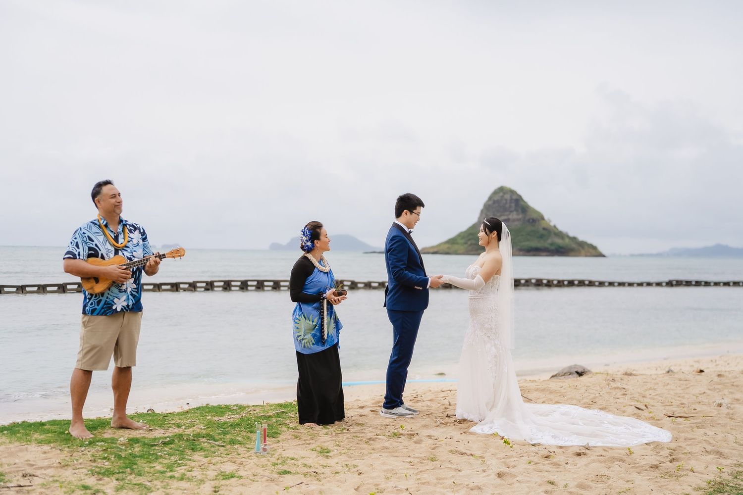 Couple exchanges vows on sandy Hawaii beach with ukulele musician and officiant, Chinaman's Hat island in background.
