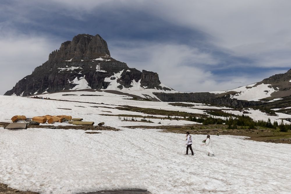 Two hikers walk across a snowy alpine meadow with a rugged mountain peak rising under cloudy skies.