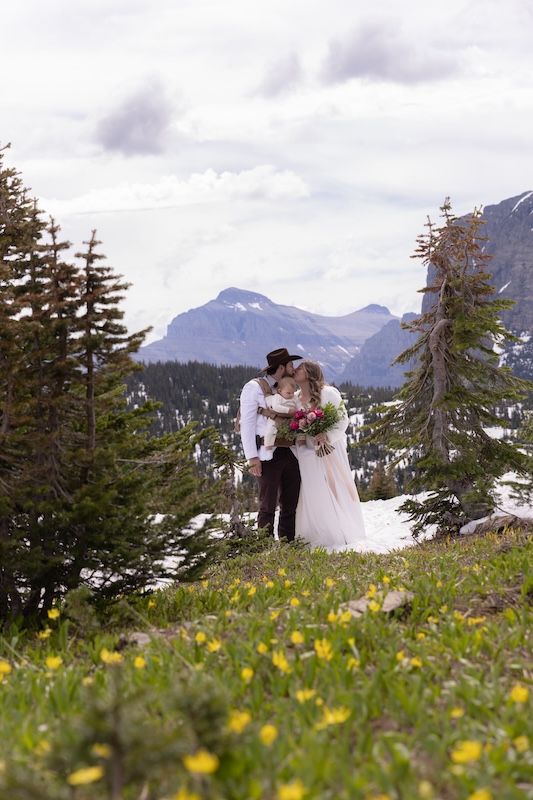A couple shares a romantic kiss in a mountain meadow with wildflowers, evergreen trees, and snowy peaks in the background.