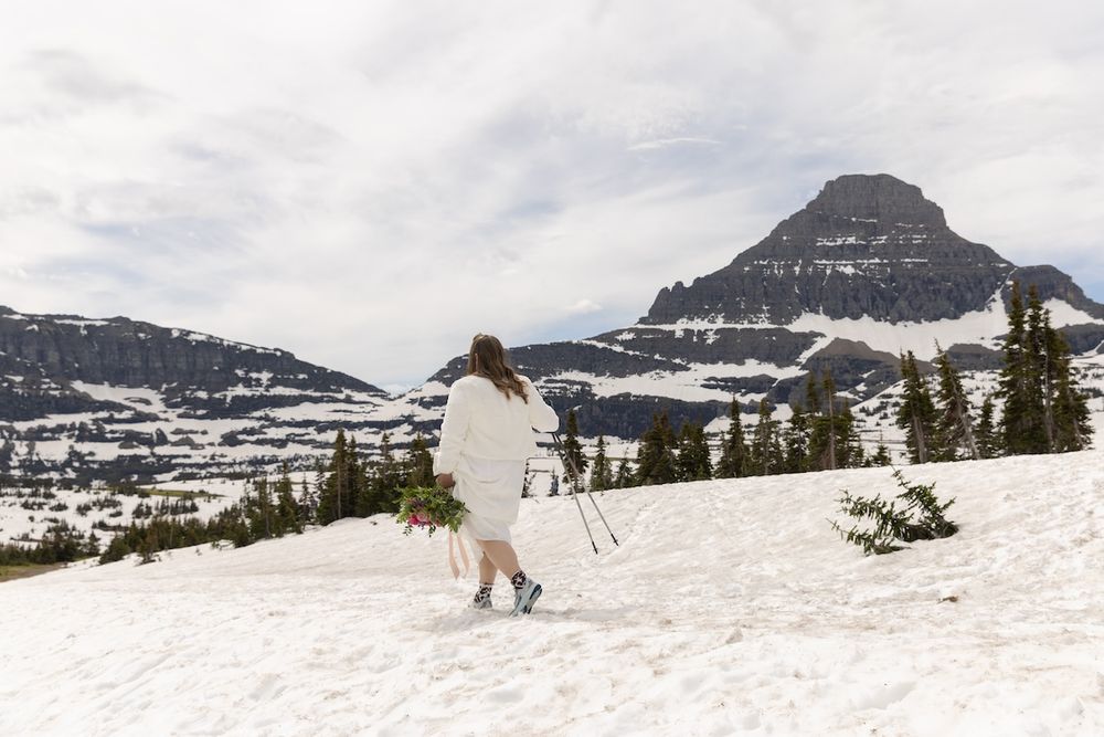 Bride in white dress walks through snow with mountain peaks and evergreen trees in background.