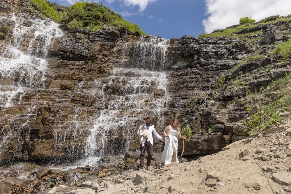 Couple in wedding attire holding hands before a cascading waterfall on a rocky mountain cliff under blue skies.