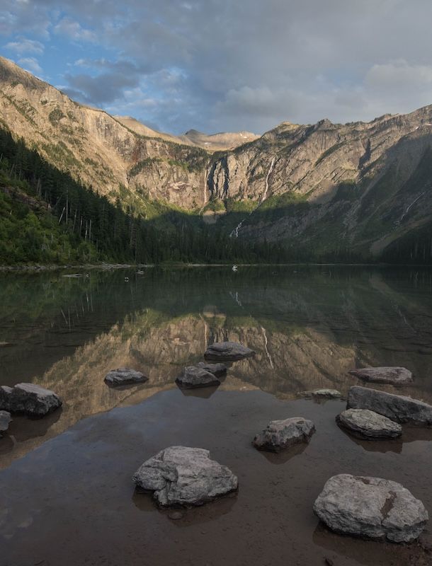 Serene mountain lake with rocky foreground, reflecting towering peaks and waterfalls under a partly cloudy sky.