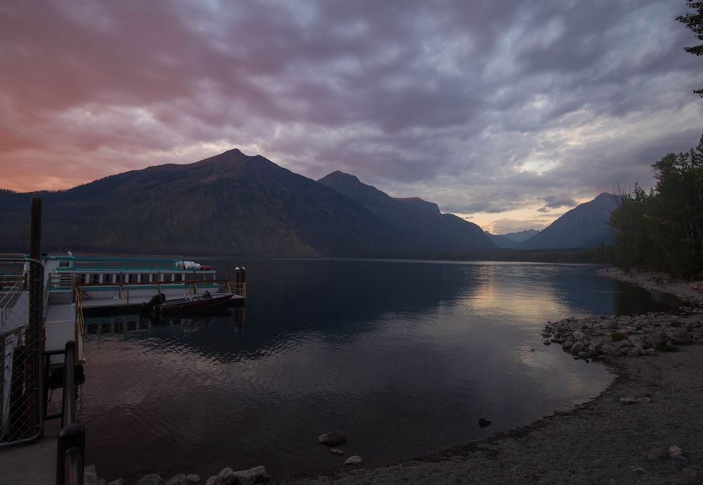 Dramatic sunset over a mountain lake with a docked boat reflecting colorful clouds in calm waters.