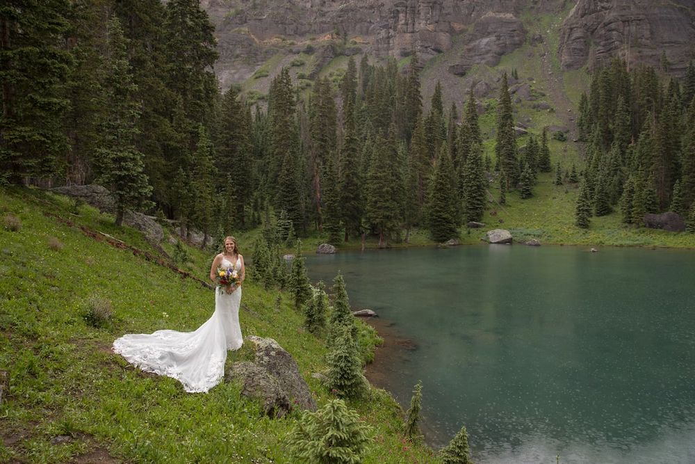 Bride in white gown stands on rocky lakeside surrounded by evergreen trees and turquoise mountain lake.