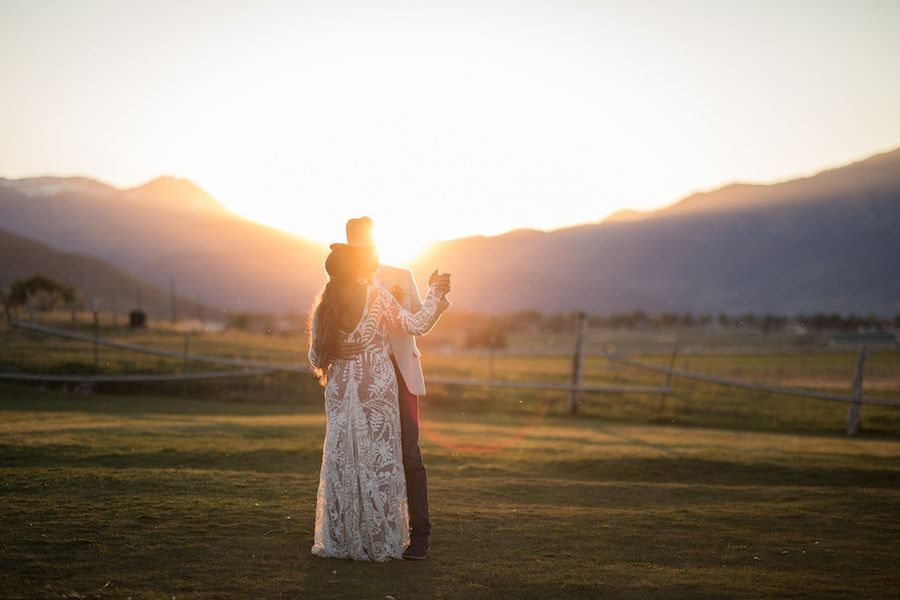 Couple embracing at golden sunset with mountain backdrop on lush green field during outdoor wedding ceremony.