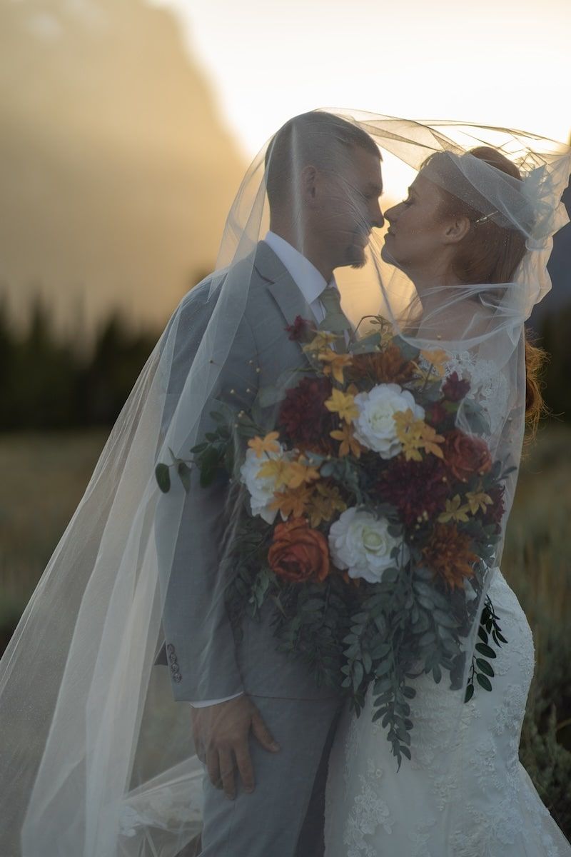 Bride and groom sharing a kiss under a veil at golden hour, holding a colorful autumn bouquet outdoors.