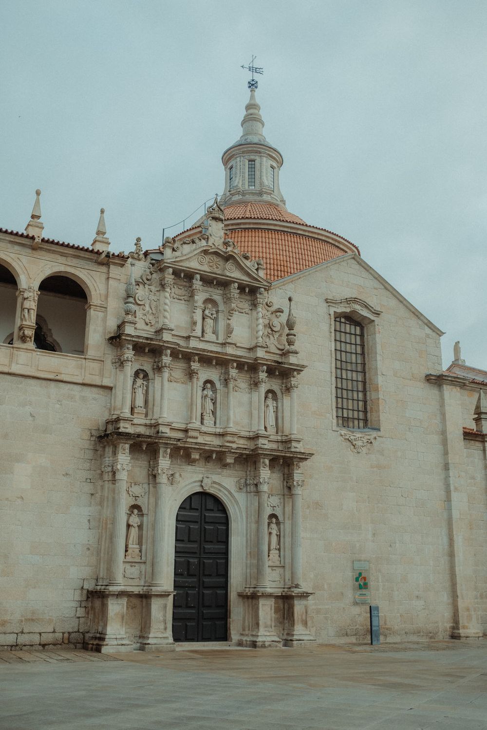 Baroque stone church facade with ornate carvings, statues, and terracotta dome under overcast sky in Spain.