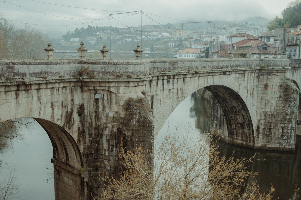 Historic stone arch bridge over a calm river in a misty European town with bare trees and old buildings in the background.
