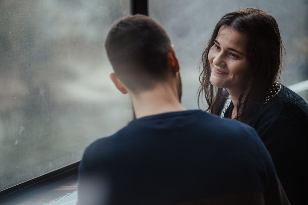 A smiling young woman with dark hair converses with a man near a rain-streaked window indoors.