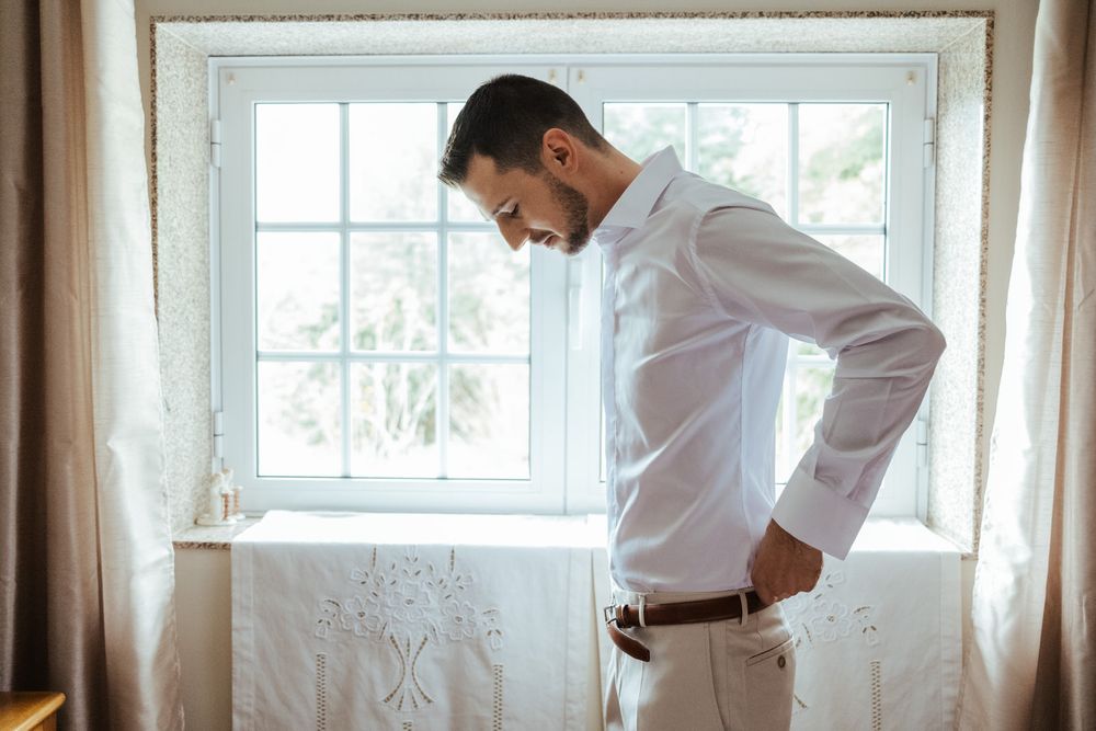 A well-dressed groom in a white shirt and tan pants stands by a bright window, looking down while adjusting his belt.