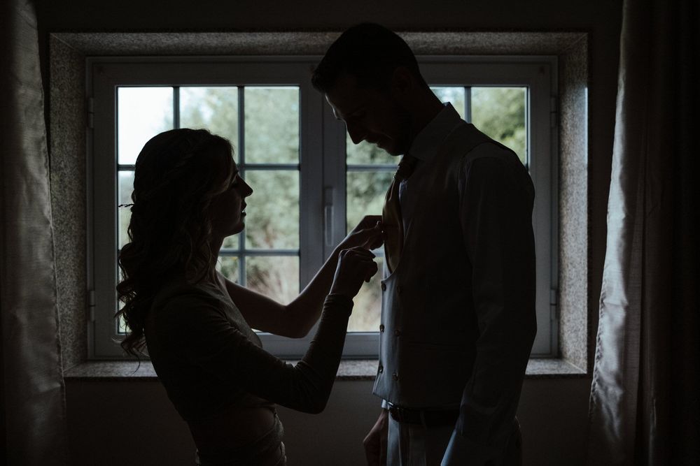 Silhouette of a bride and groom standing by a window, sharing an intimate moment as she adjusts his tie.