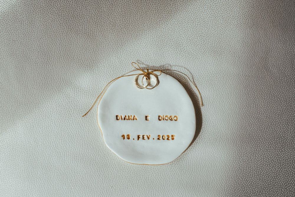 White ceramic wedding ring dish with gold text reading DIANA E DIOGO 25.FEV.2023, tied with a bow.