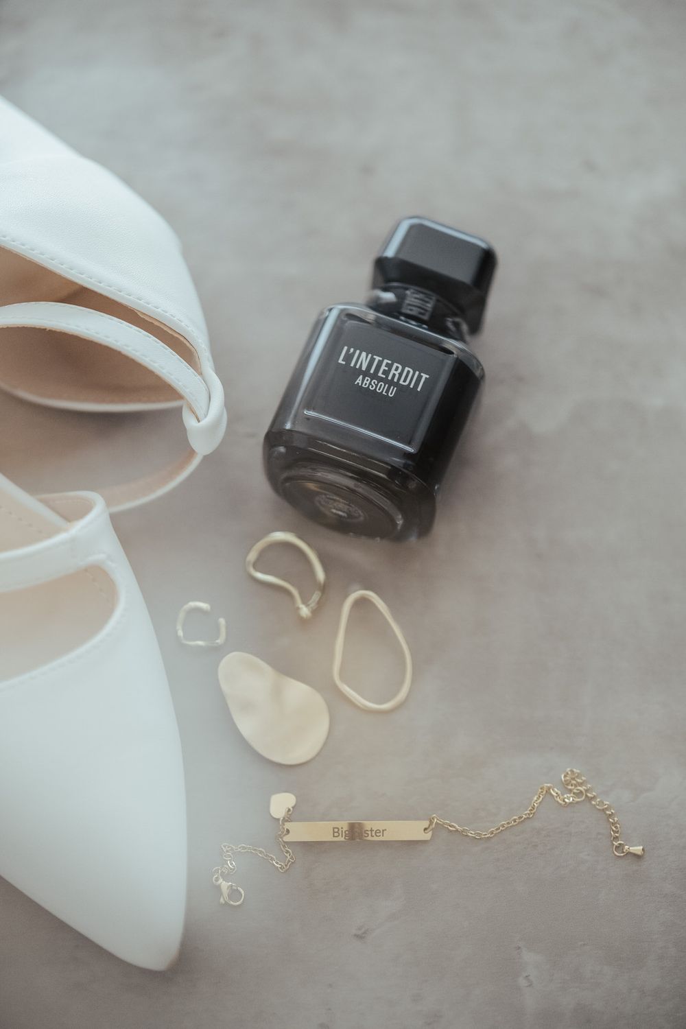 Bridal flat lay with white heels, L'Interdit Absolu perfume, gold bracelet, and shoe inserts on gray surface.