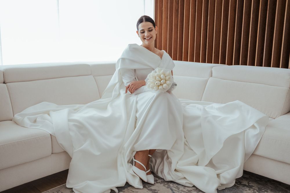Elegant bride in white wedding gown with cape sleeves sits on cream sofa holding white floral bouquet.