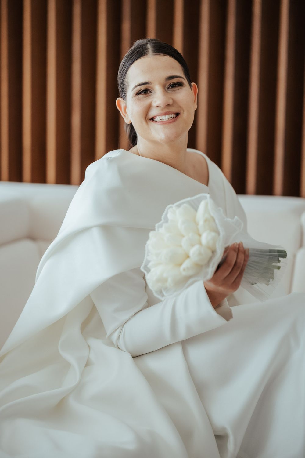 Smiling bride in a white wedding gown holding a white tulip bouquet while seated.