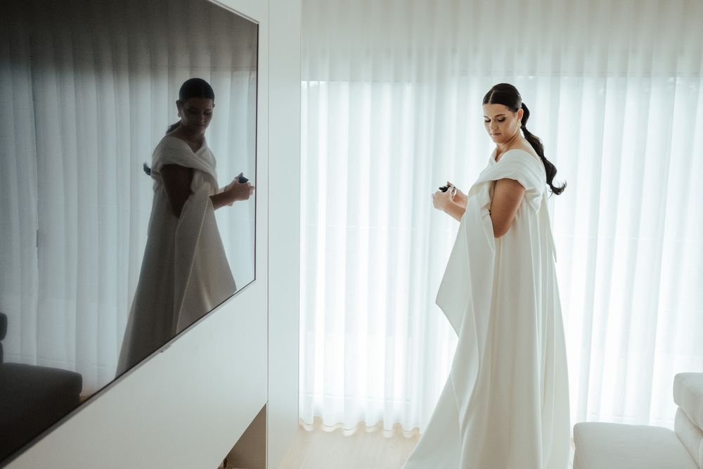 Elegant bride in a white off-shoulder gown admiring her reflection in a mirror.