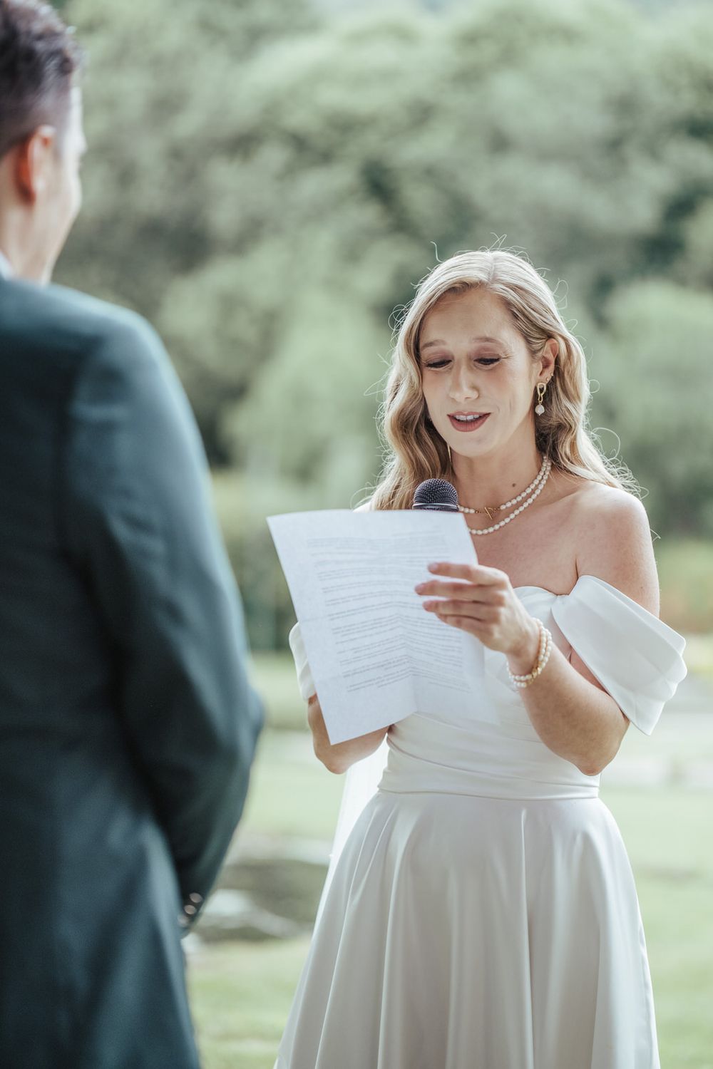 Bride in white off-shoulder dress reads wedding vows from paper during outdoor ceremony.