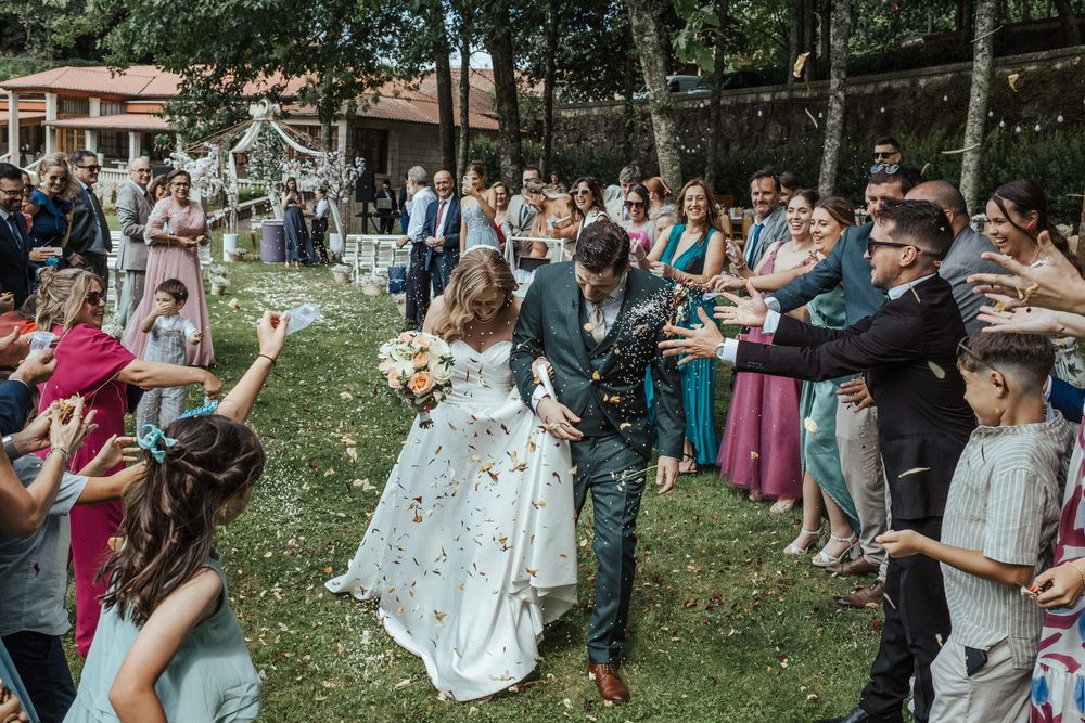 Bride and groom kiss while guests toss confetti during outdoor wedding ceremony on lush green lawn.