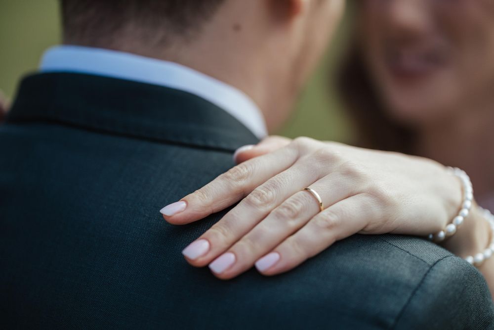 Close-up of a bride's hand with a gold wedding ring resting on the groom's dark suit jacket shoulder.