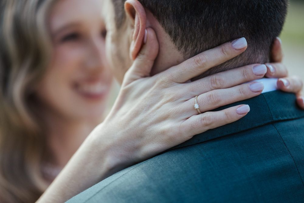 Close-up of a woman's hand with a gold engagement ring resting on a man's shoulder as they embrace.