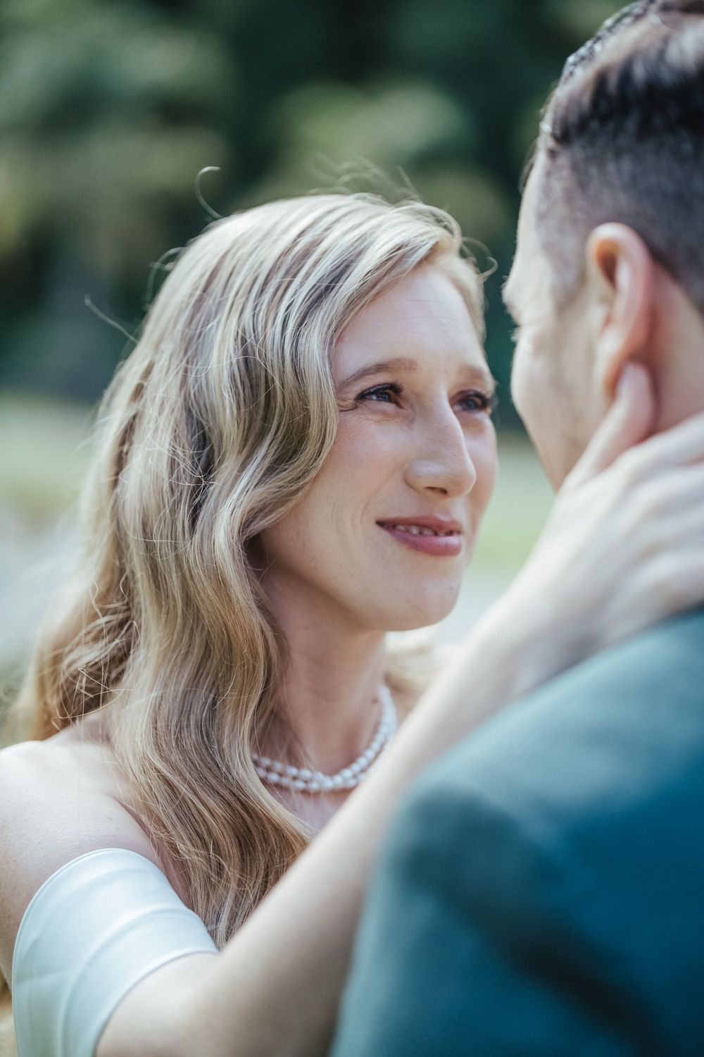Bride in white dress and pearl necklace gazes lovingly at groom during outdoor wedding ceremony.