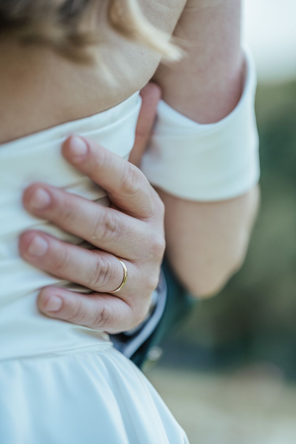Close-up of groom's hand with gold wedding band embracing bride in white dress during wedding ceremony.